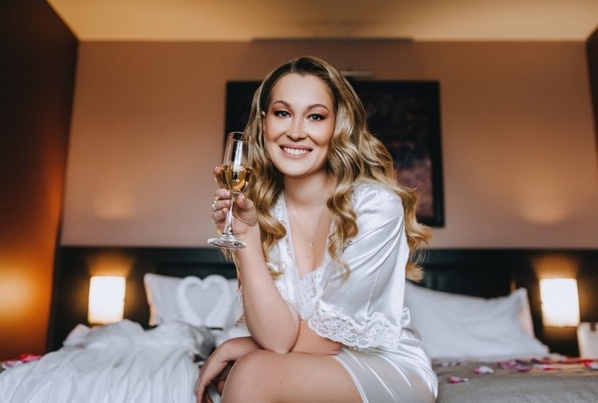 A bride in a robe with a glass of champagne on a bed | Skylum Blog